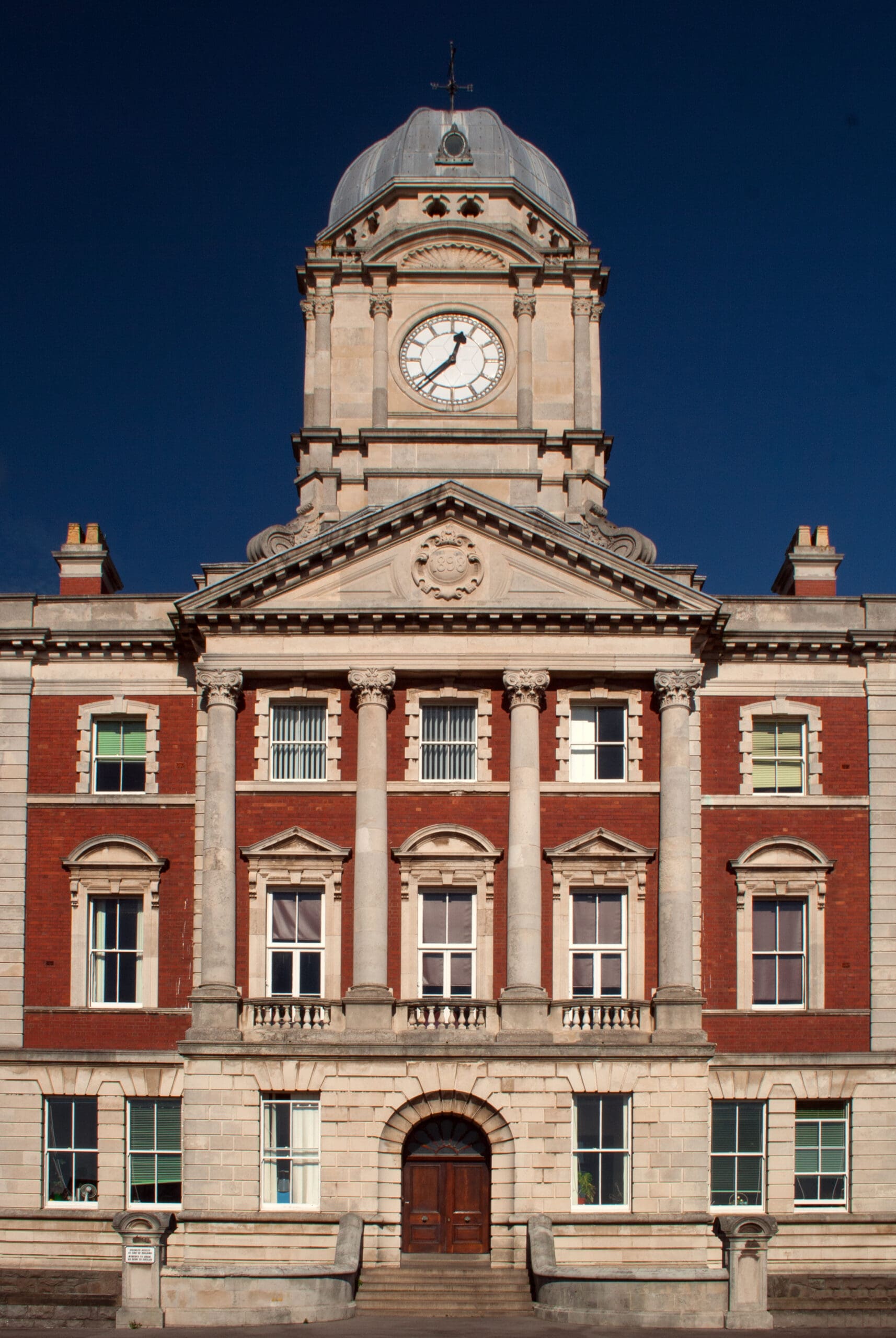 A large, ornate building with a clock tower at the center. The facade is a mix of red brick and stone, with tall columns and arched windows. The clock is prominent and shows 5 o’clock. The building has a historical architectural style.