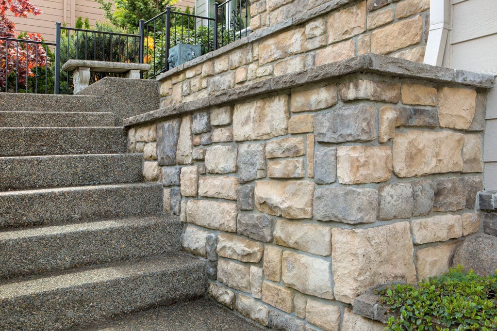 A close-up view of an exterior stone wall and staircase made of speckled concrete leading up to a house entrance. Adjacent to the stairs is a short black metal railing, and surrounding the area is greenery and a garden with a stone bench.