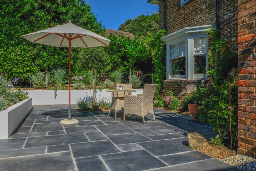 A sunny patio features a slate tile floor with a round table and two wicker chairs shaded by a large white umbrella. Surrounding the space are lush green plants and shrubs, and a bay window on the brick house overlooks the garden.