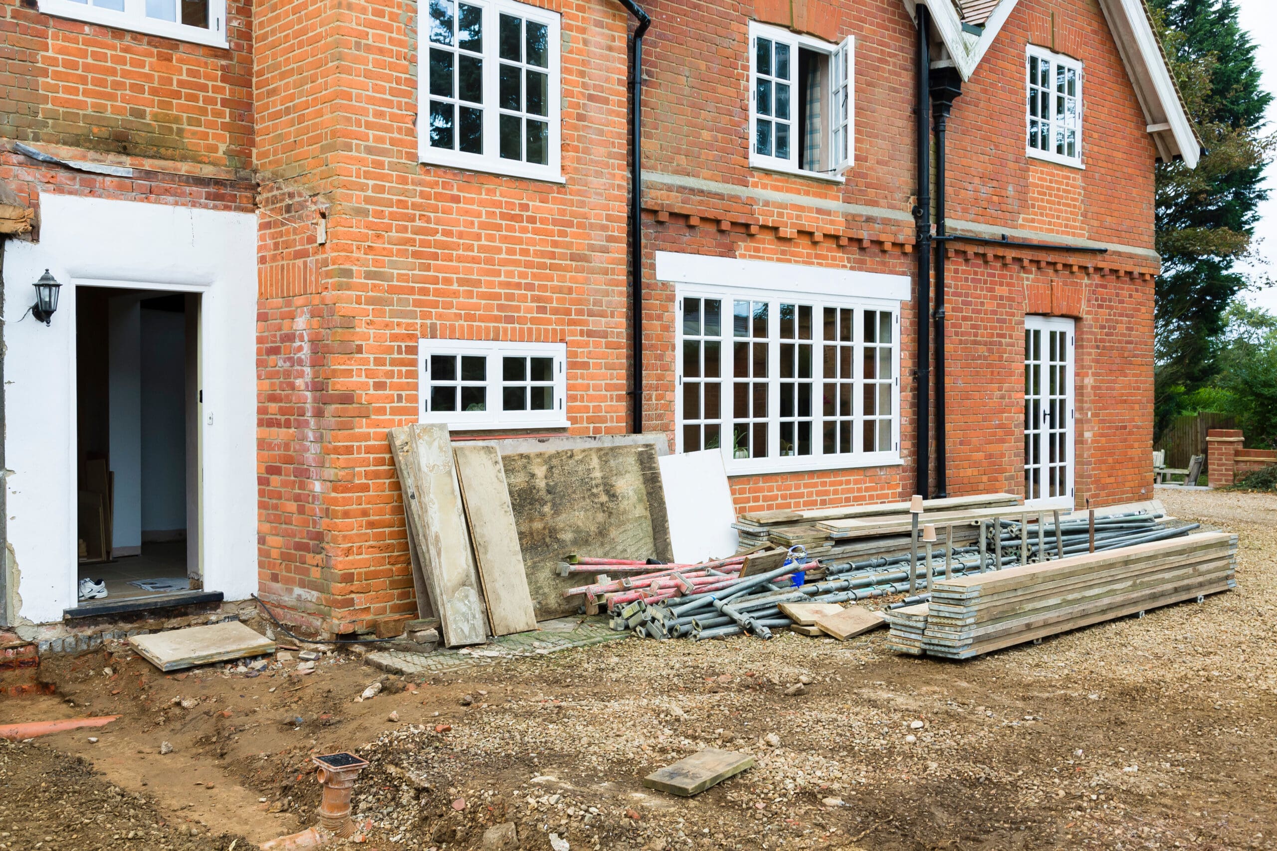 A construction site in front of a partially completed brick house with large windows. Scaffolding, wooden boards, and various other construction materials are scattered around. The ground is covered in gravel, and a tree is visible in the background.