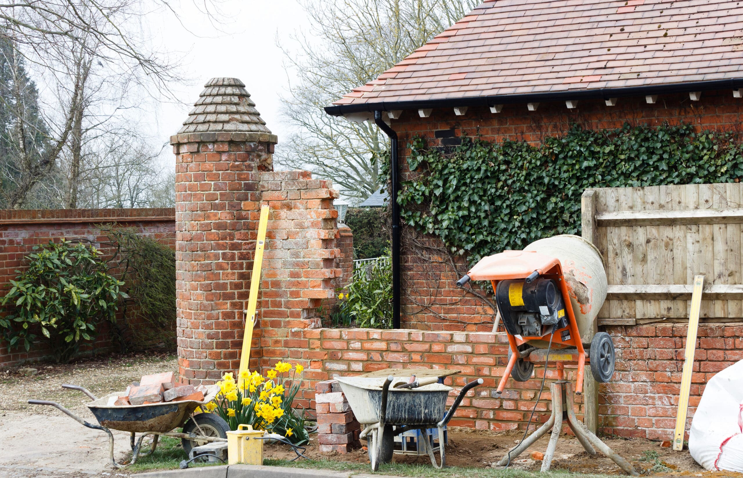 A construction site with brick wall repairs in progress. A partially rebuilt brick wall features a chimney-like structure. A cement mixer, wheelbarrows, bricks, ladders, and tools are scattered around. Yellow flowers bloom near the wall, and a building with ivy is in the background.