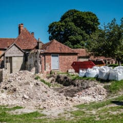 A red-brick house with a sloped roof is under renovation. Construction materials, including white bags and a red dumpster, are scattered around the site. A large tree and a clear blue sky are in the background, while the foreground shows piles of rubble.