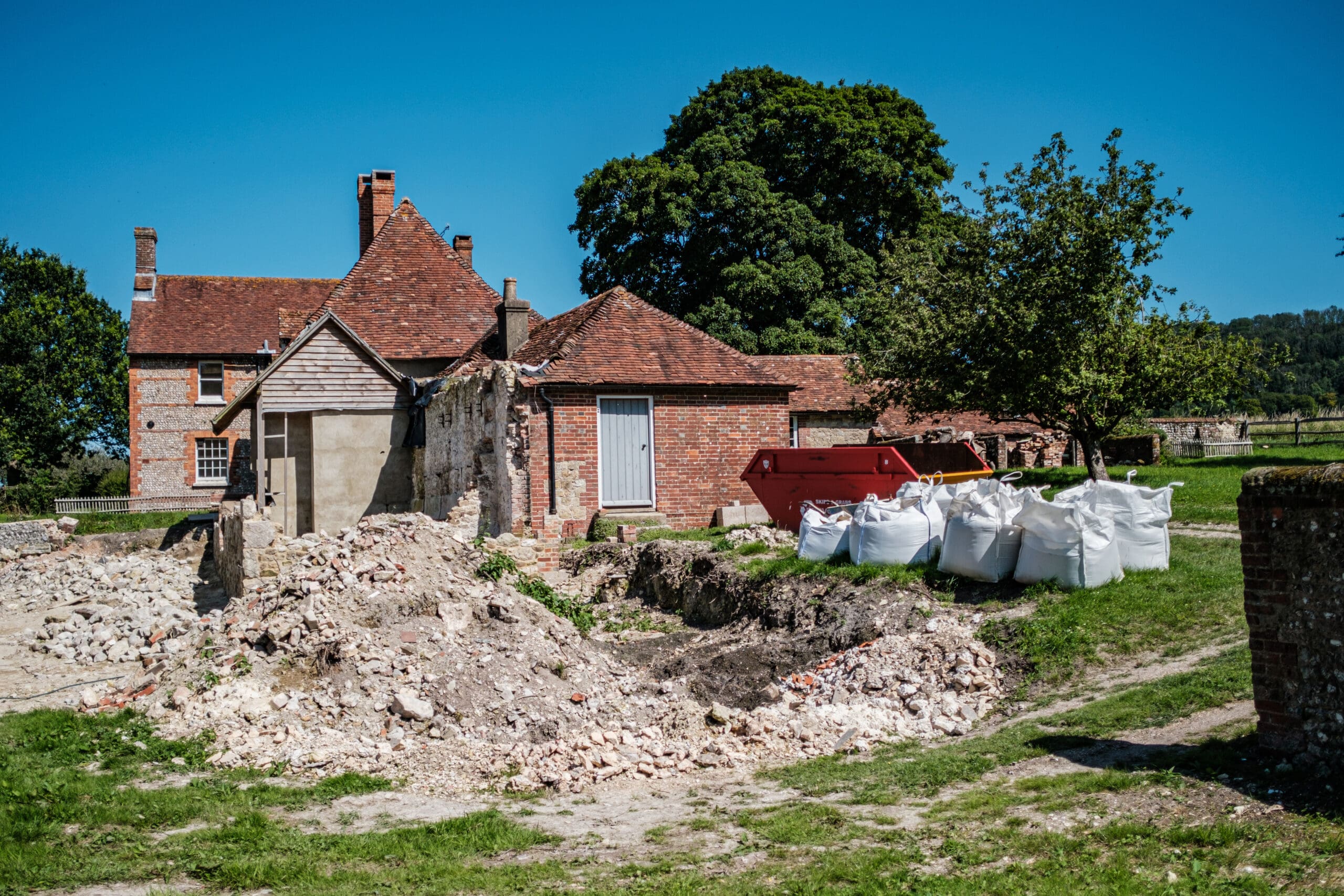 A red-brick house with a sloped roof is under renovation. Construction materials, including white bags and a red dumpster, are scattered around the site. A large tree and a clear blue sky are in the background, while the foreground shows piles of rubble.