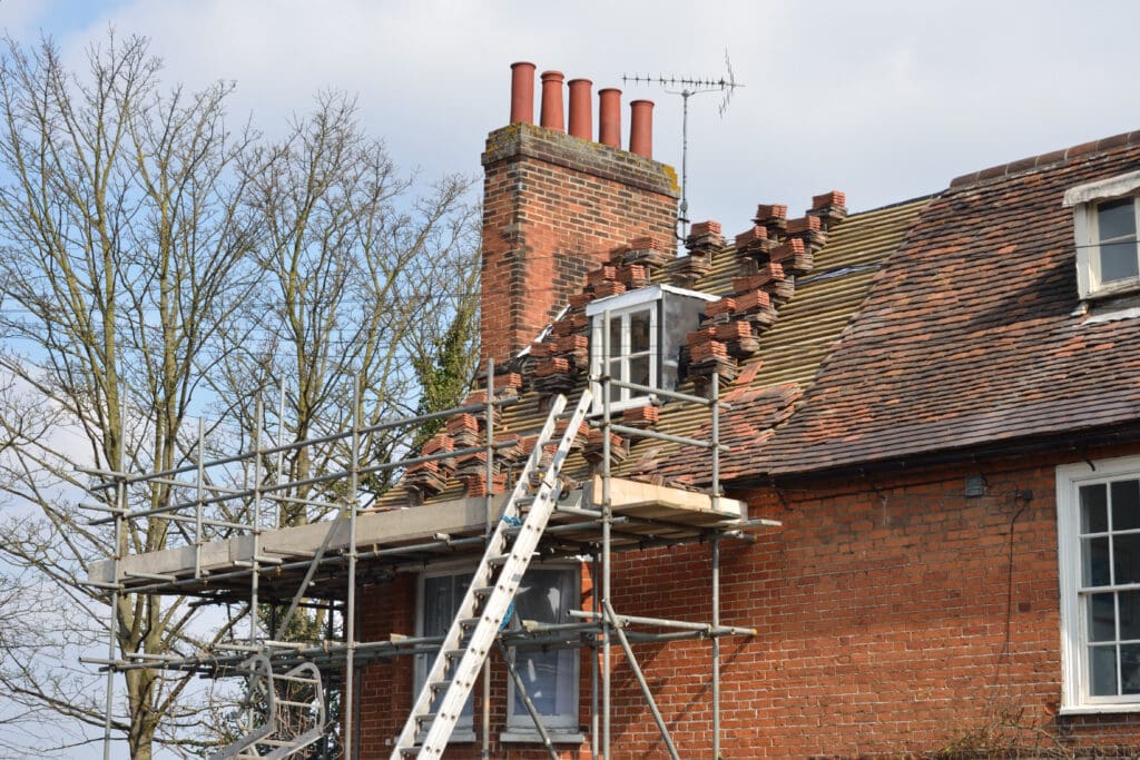 A brick house with a construction scaffold on the side. The roof is partially removed as part of ongoing repairs, with exposed wooden beams visible. Ladders are propped against the scaffold, and bare trees are in the background. A TV antenna is on the chimney.