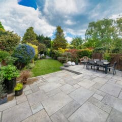 A spacious, well-maintained backyard features a large stone patio with an outdoor dining table and chairs. Surrounding the patio are lush, vibrant plants and trees, leading to a neatly manicured, green lawn. The sky above is partly cloudy.