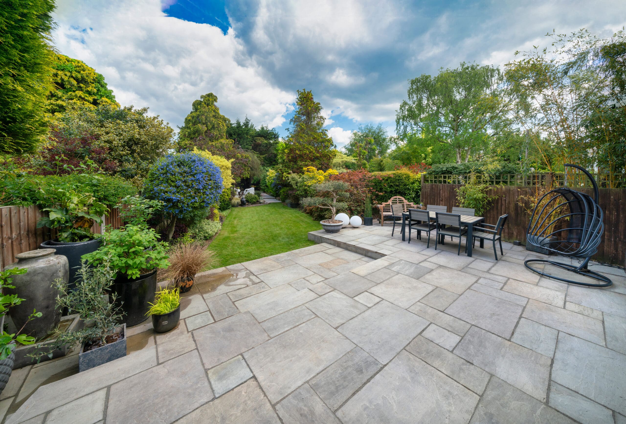 A spacious, well-maintained backyard features a large stone patio with an outdoor dining table and chairs. Surrounding the patio are lush, vibrant plants and trees, leading to a neatly manicured, green lawn. The sky above is partly cloudy.