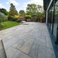 A spacious backyard with a large stone patio adjacent to a house with sliding glass doors on the right. The patio steps down to a green lawn surrounded by a variety of lush plants, trees, and colorful flowers. The sky is clear and bright.