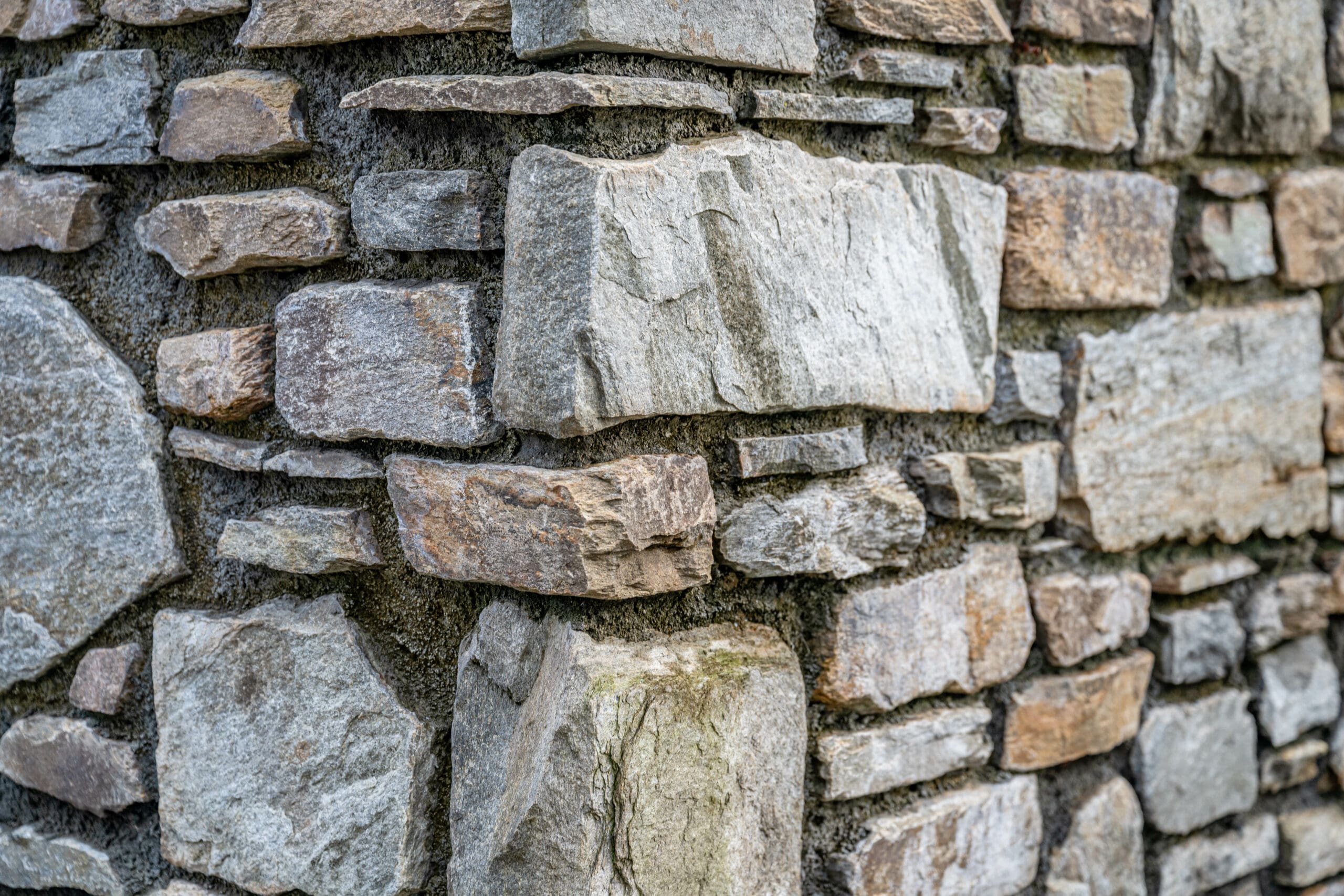 Close-up of a rustic stone wall made of variously shaped and sized stones. The stones are predominantly gray with hints of brown and beige, and are laid in an irregular pattern with visible mortar joining them. The texture is rough and weathered.