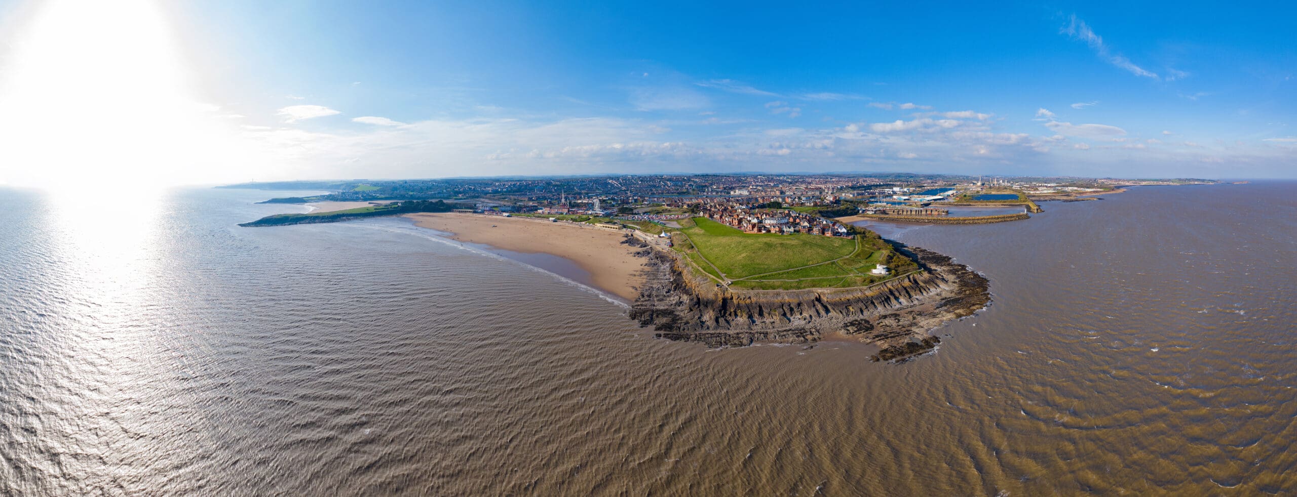 Aerial view of a coastal town with a large sandy beach, rocky cliffs, and grassy open spaces extending into the water. The town is situated along the coastline, with buildings and structures visible. The sea is calm, and the sky is clear and blue.