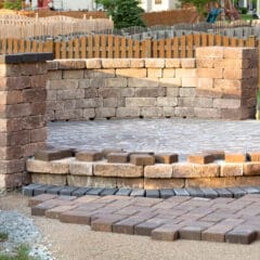 A partially completed circular patio area made of interlocking pavers with a low stone wall enclosing a section. The construction site appears to be in a residential backyard with a wooden fence and lawn in the background.