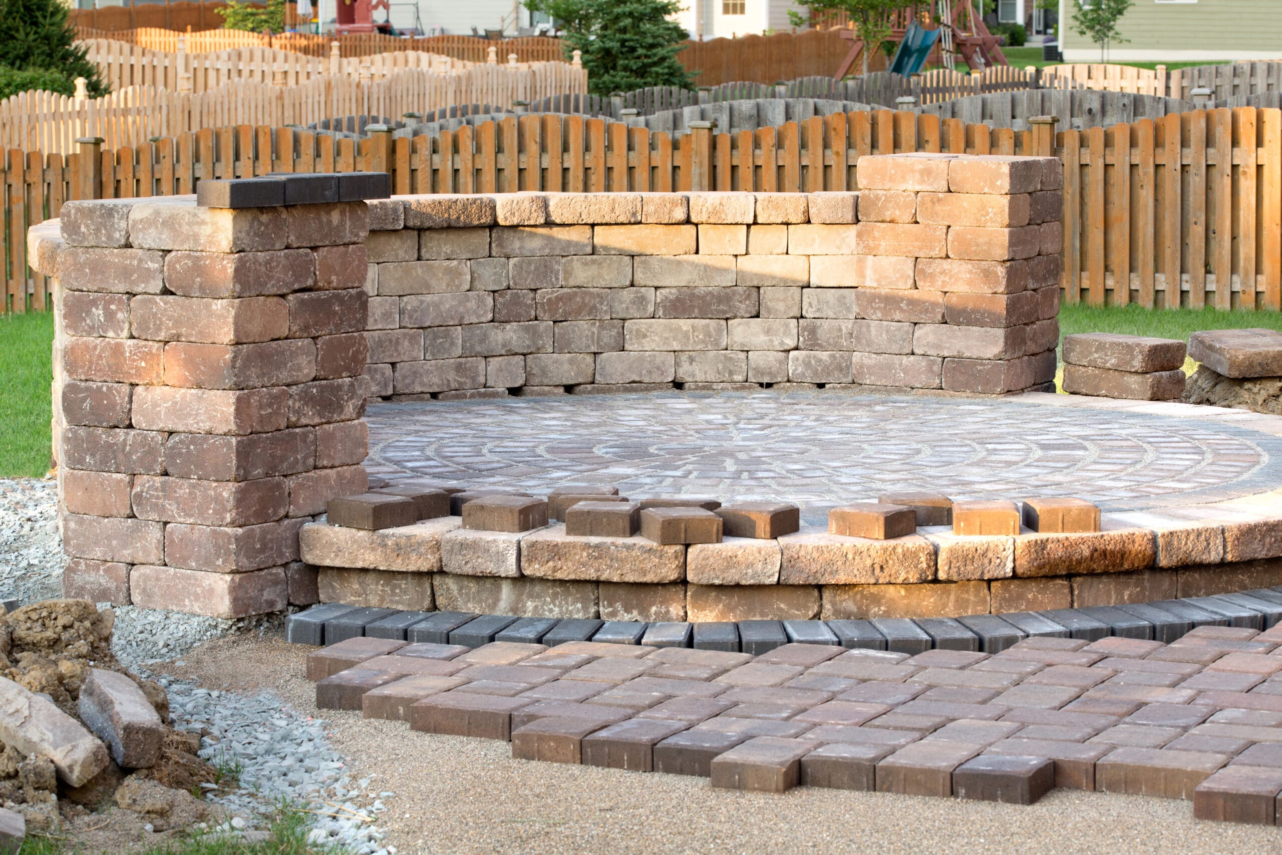 A partially completed circular patio area made of interlocking pavers with a low stone wall enclosing a section. The construction site appears to be in a residential backyard with a wooden fence and lawn in the background.