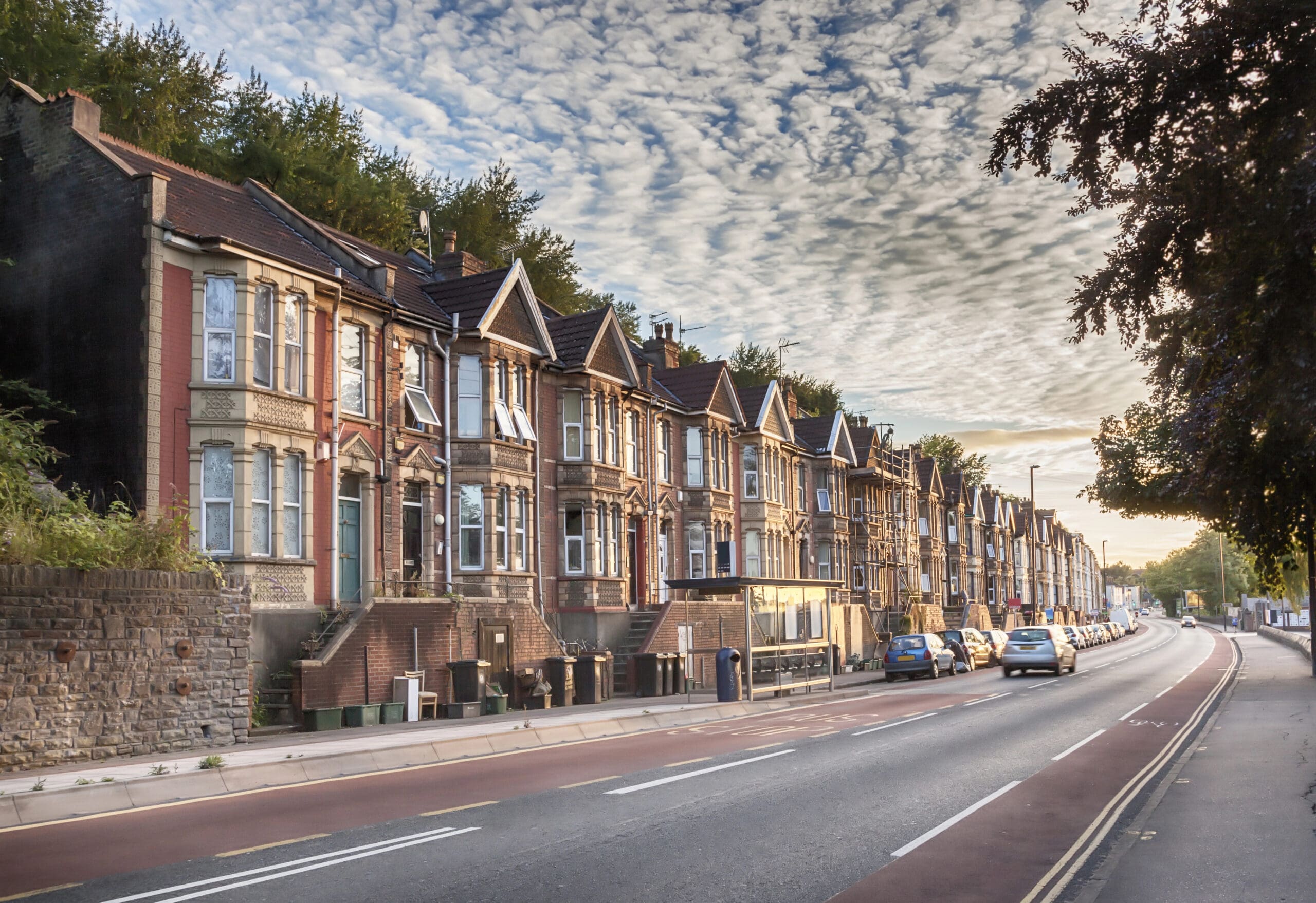 A row of picturesque, Victorian-style terraced houses lines a quiet street under a partly cloudy sky. The houses feature bay windows and decorative brickwork. Several parked cars are visible along the street, which is empty of traffic. Trees can be seen in the background.