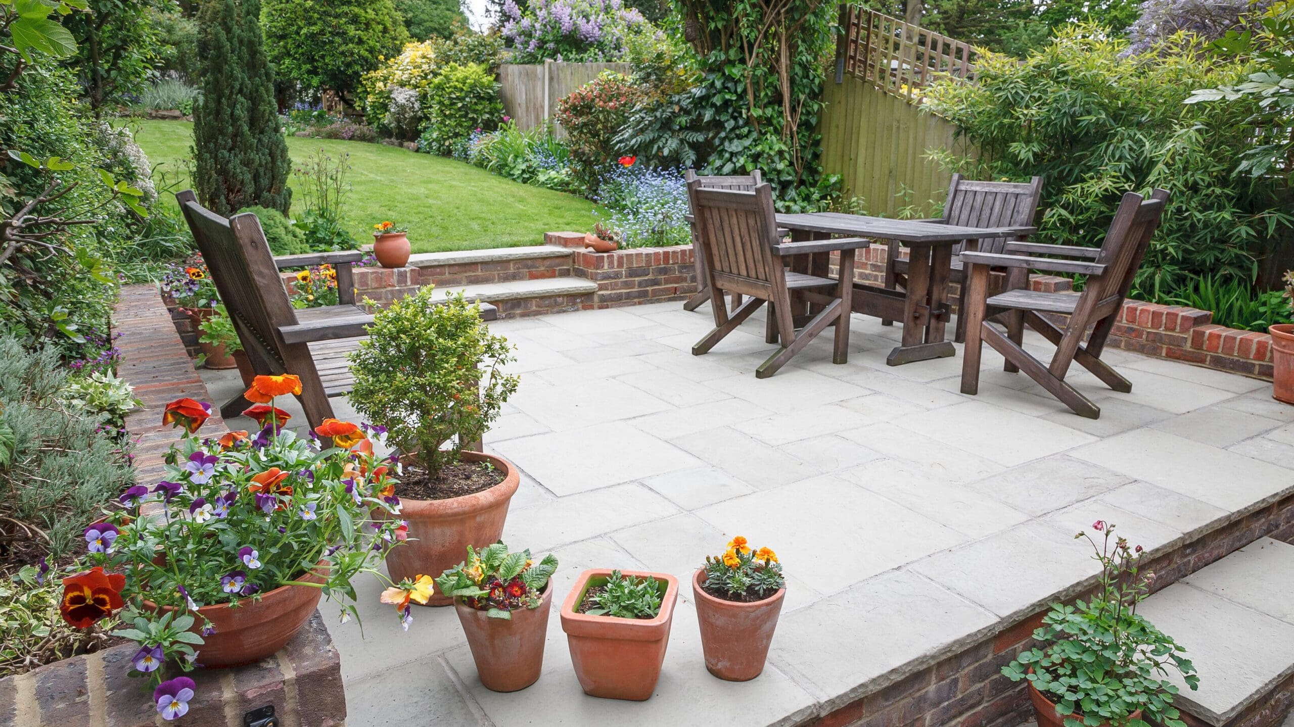 A serene garden patio with wooden chairs and a table on a tiled surface. Potted flowers in various colors are arranged around the patio. The lush green garden area with trees, shrubs, and a lawn extends in the background, providing a tranquil setting.