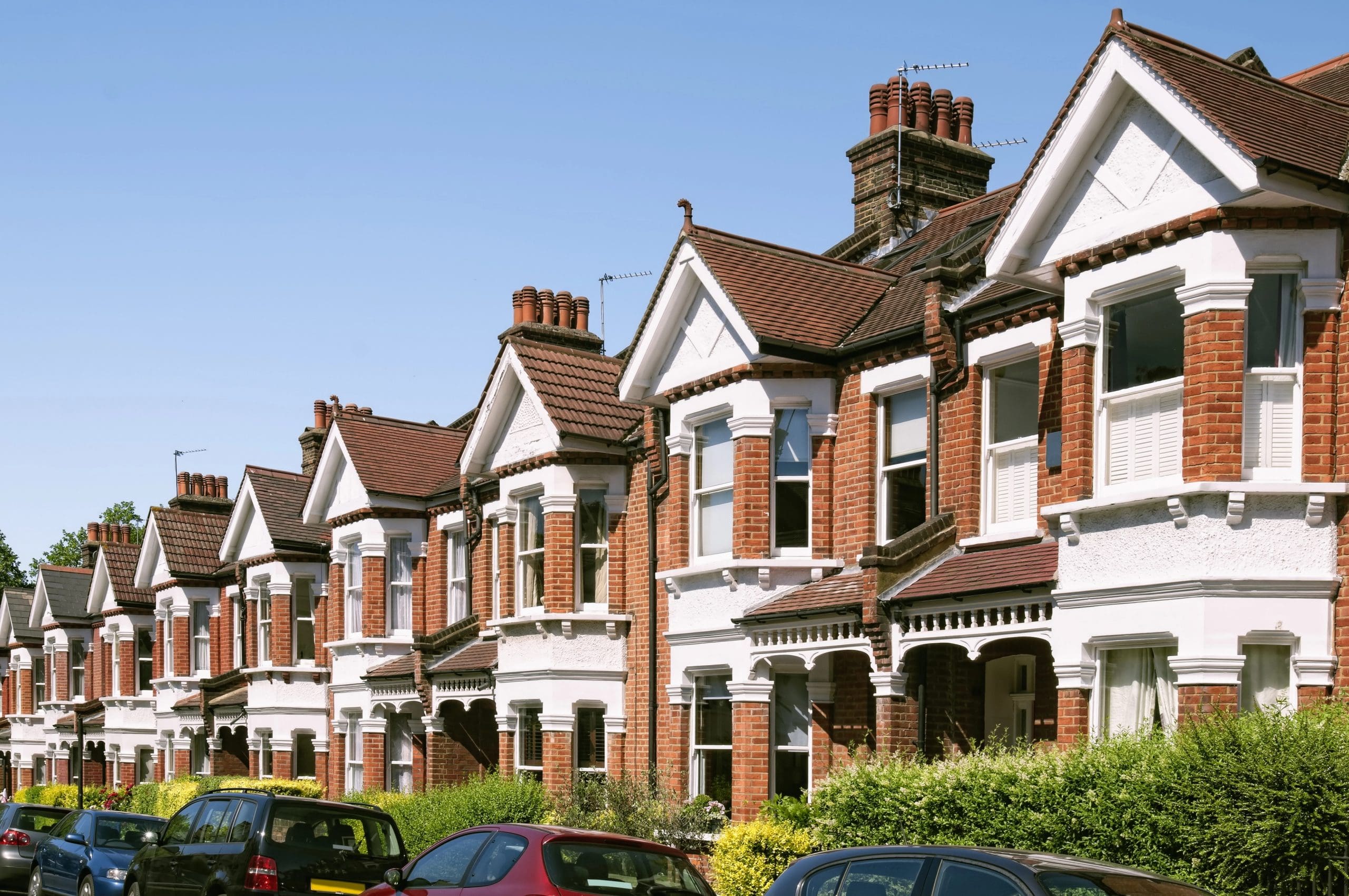 A row of traditional red-brick terraced houses with white window frames and gabled roofs under a clear blue sky. Cars are parked along the street in front of neatly trimmed hedges and gardens.