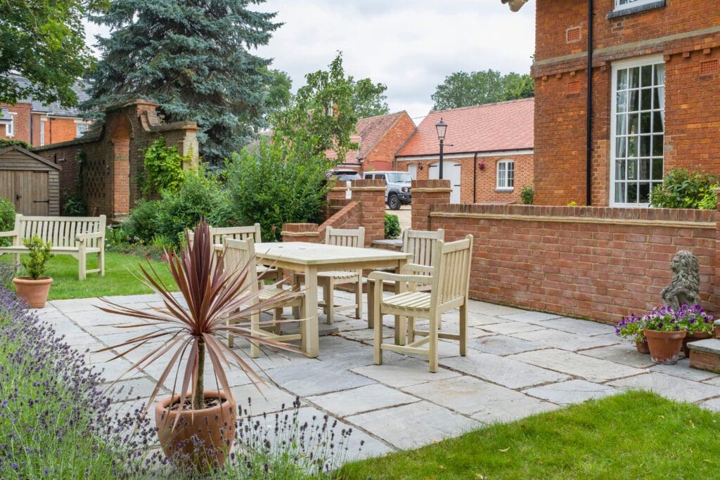 A stone patio with a wooden table and six chairs is surrounded by a garden. There are potted plants and a trimmed lawn. In the background, there are brick buildings and a parked white vehicle. A large evergreen tree and various shrubs are visible.