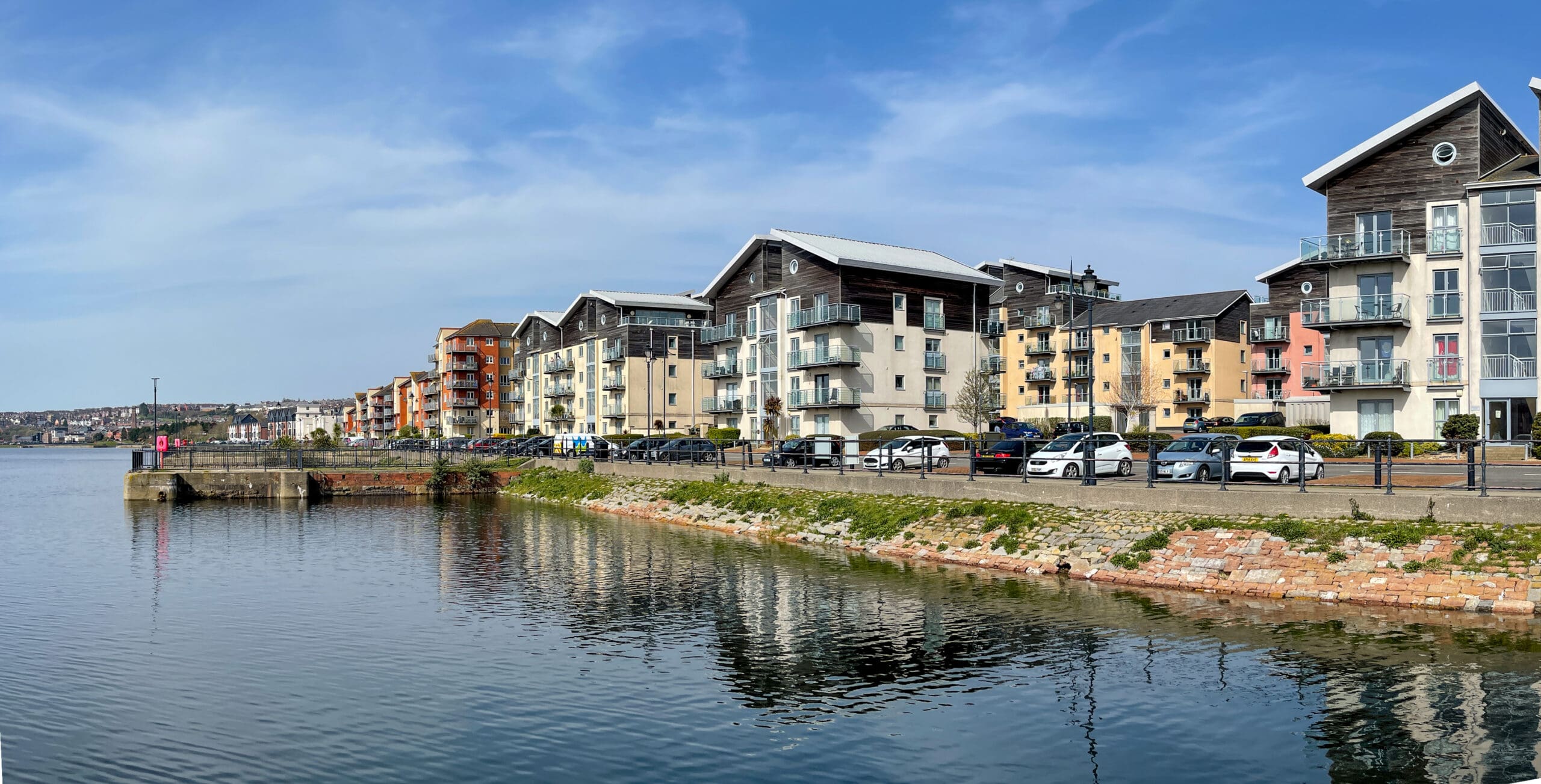 A waterfront view of modern apartment buildings with balconies, lined up along the shore under a blue sky. Parked cars are visible along the street in front of the buildings. The calm water reflects parts of the buildings and a stone embankment.