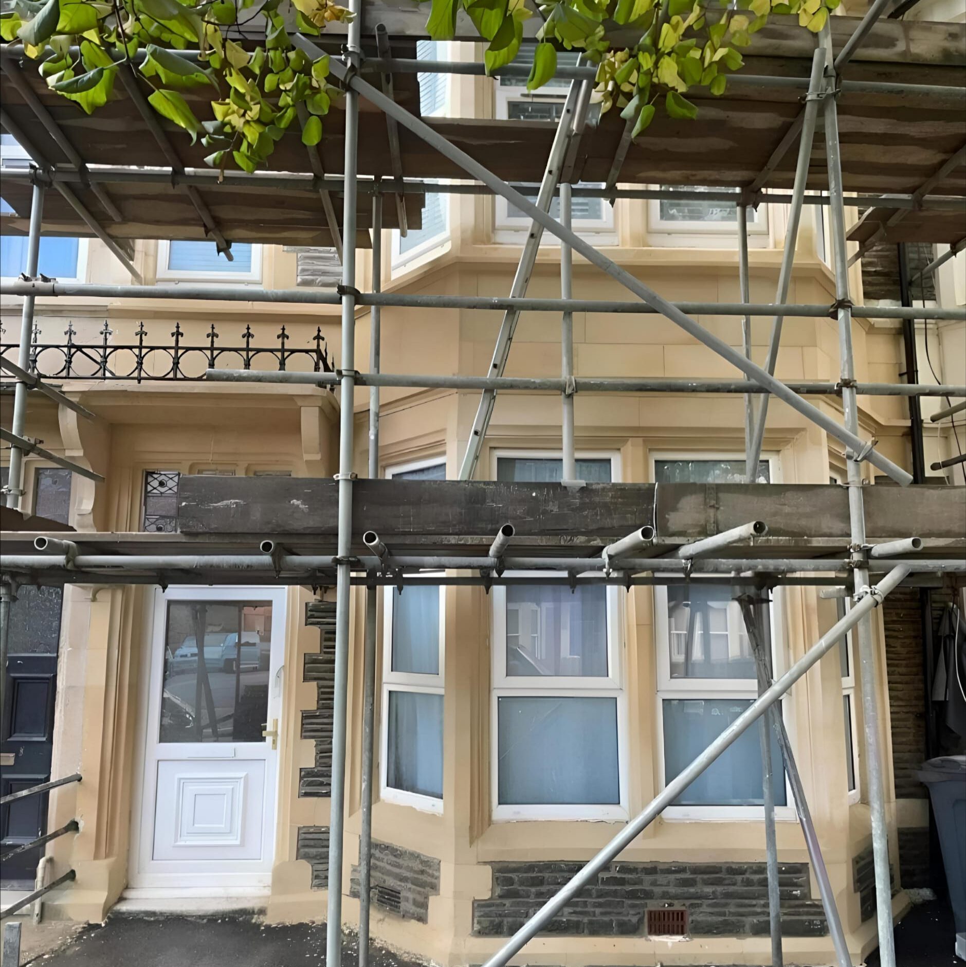 A two-story house with beige walls and large bay windows is surrounded by metal scaffolding. The entrance door is white with a small window above, and there are green leaves hanging from the top left corner.