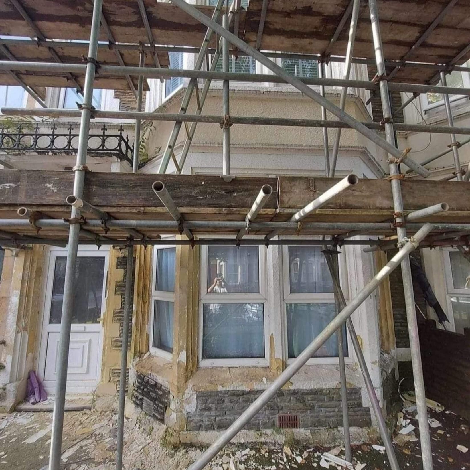 A building under construction with scaffolding surrounding the exterior. The windows and door of the lower level are visible, with peeling paint and debris scattered on the ground. The structure appears to be undergoing renovation or restoration.