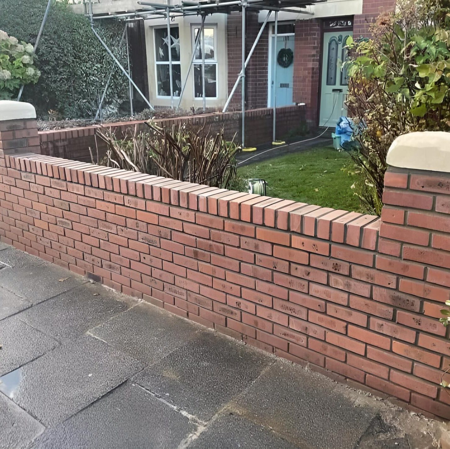 A red brick wall lines the front of a house with scaffolding on the exterior. The house has white-framed windows and a green front door with a wreath. There is a garden with trimmed grass and some plants behind the wall. The sidewalk in front appears wet.