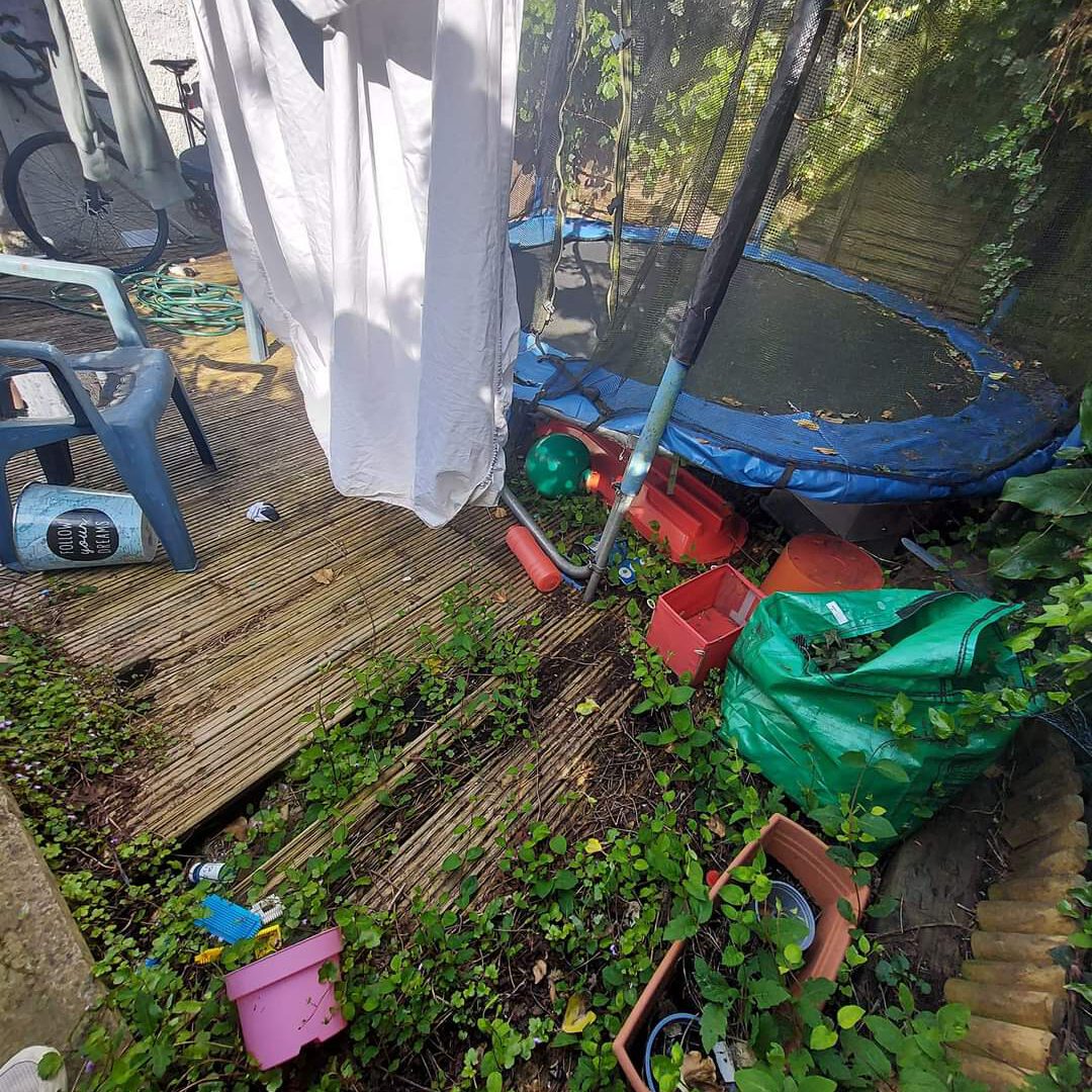 A cluttered backyard with a wooden deck, overgrown plants, and a blue trampoline surrounded by netting. Scattered items include chairs, a watering can, garden tools, a green tarp, toys, and planters. A white sheet hangs, partially covering the space.