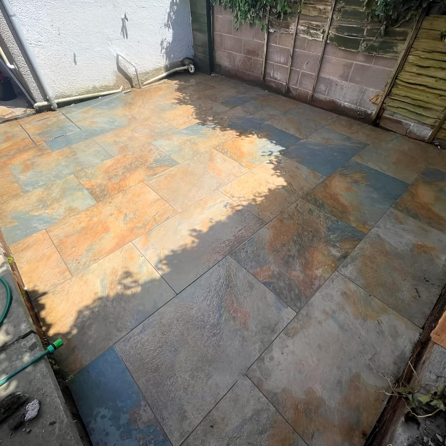 A backyard patio with newly installed multicolored slate tiles, featuring various shades of orange, green, and grey. The area is bordered by an old concrete block wall and a wooden fence with climbing plants. A garden hose is in the lower left corner.