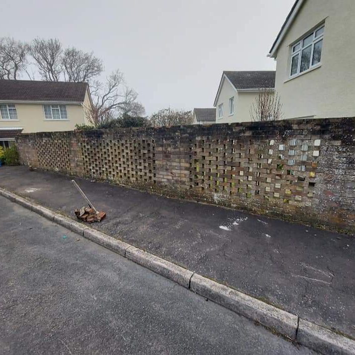 A sidewalk with a gutter broom and scattered debris. The pavement runs alongside a perforated brick wall, with several houses visible in the background. The sky is overcast, and bare trees can be seen beyond the wall.