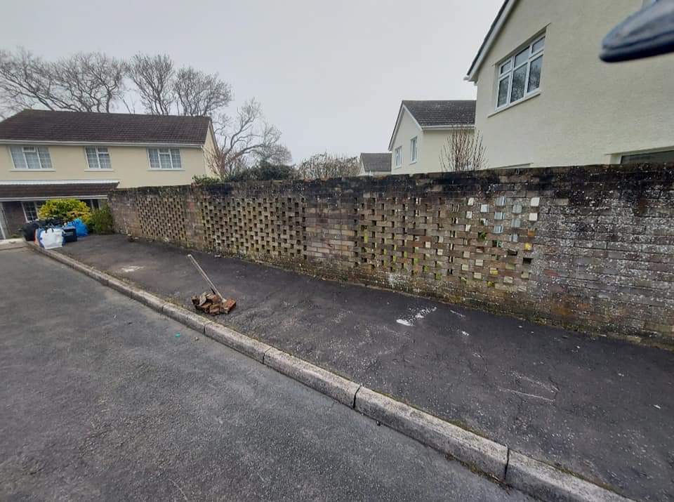 A quiet residential street with a brick wall lining the right side. In front of the wall, there is a broom and a pile of debris on the asphalt sidewalk. To the left, houses with beige exteriors and brown roofs are visible. The street is clean and empty.