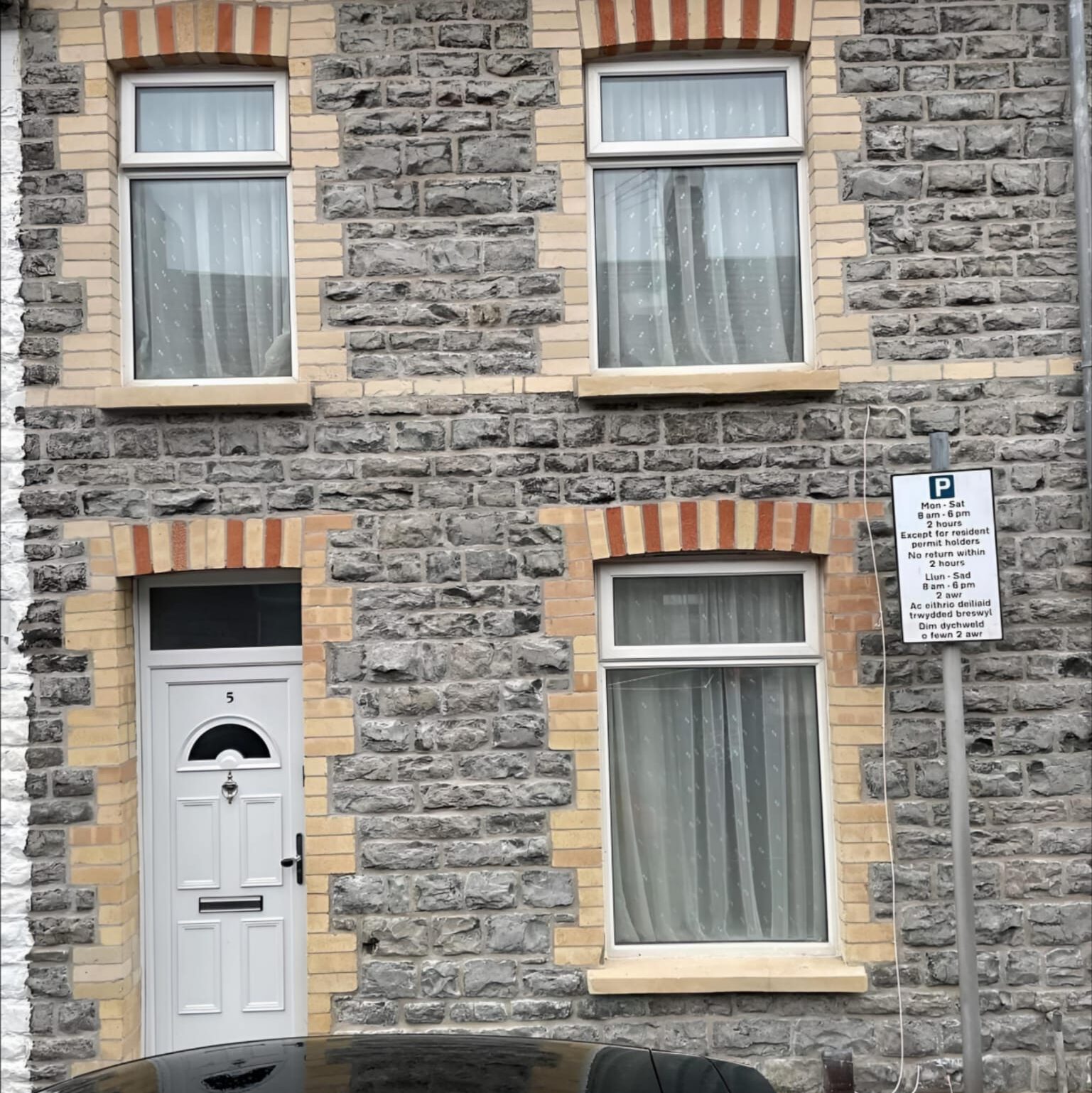 A stone-faced house with three rectangular windows, two upstairs and one downstairs, each with white curtains. A white door with a small window is next to the lower window. A "No Parking" sign is on a post by the house. Part of a car hood is visible at the bottom.