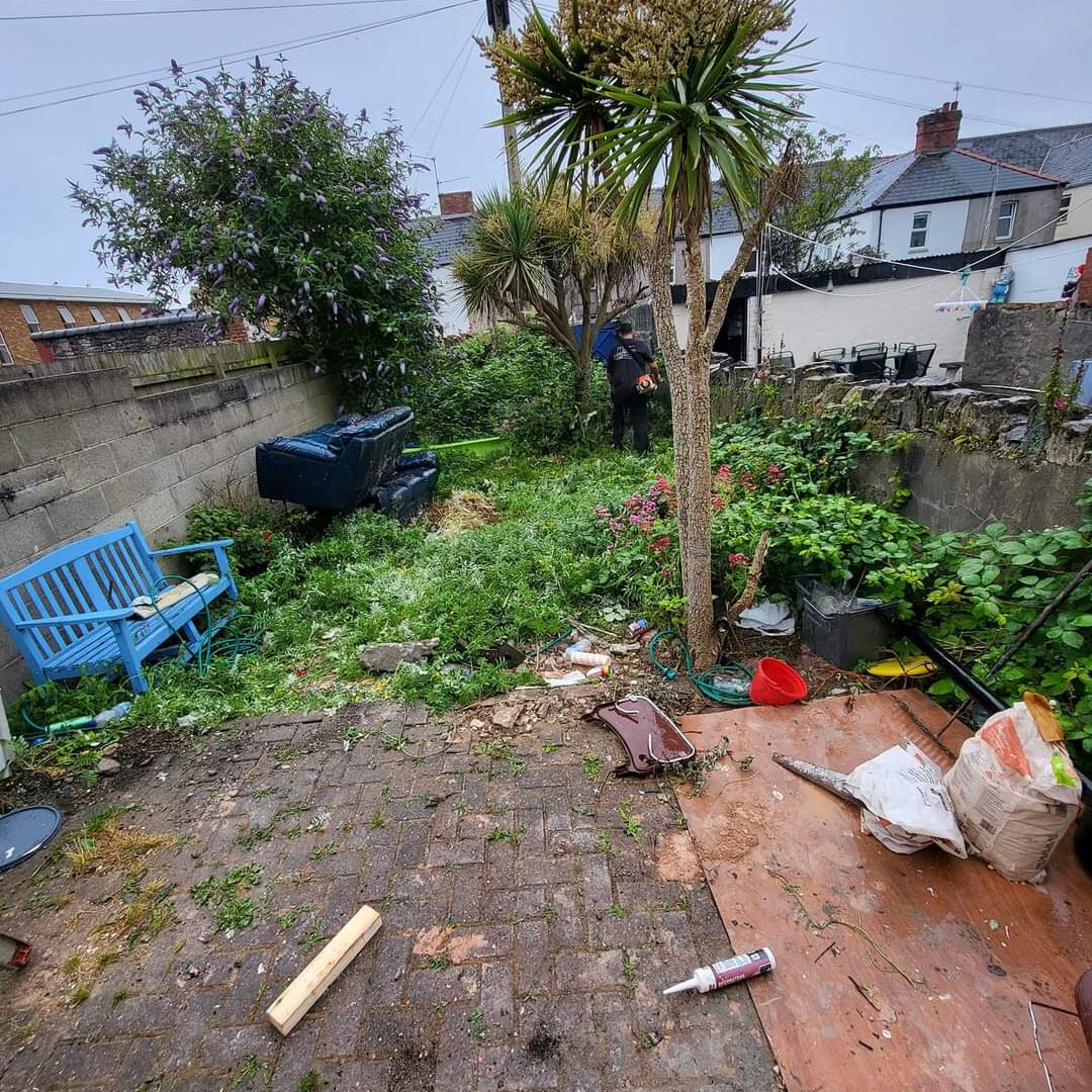 A cluttered backyard with overgrown plants and debris scattered around. There's a blue bench on the left, plastic containers, and various garden tools scattered on the ground. A person in dark clothing is standing in the distance near the fence.