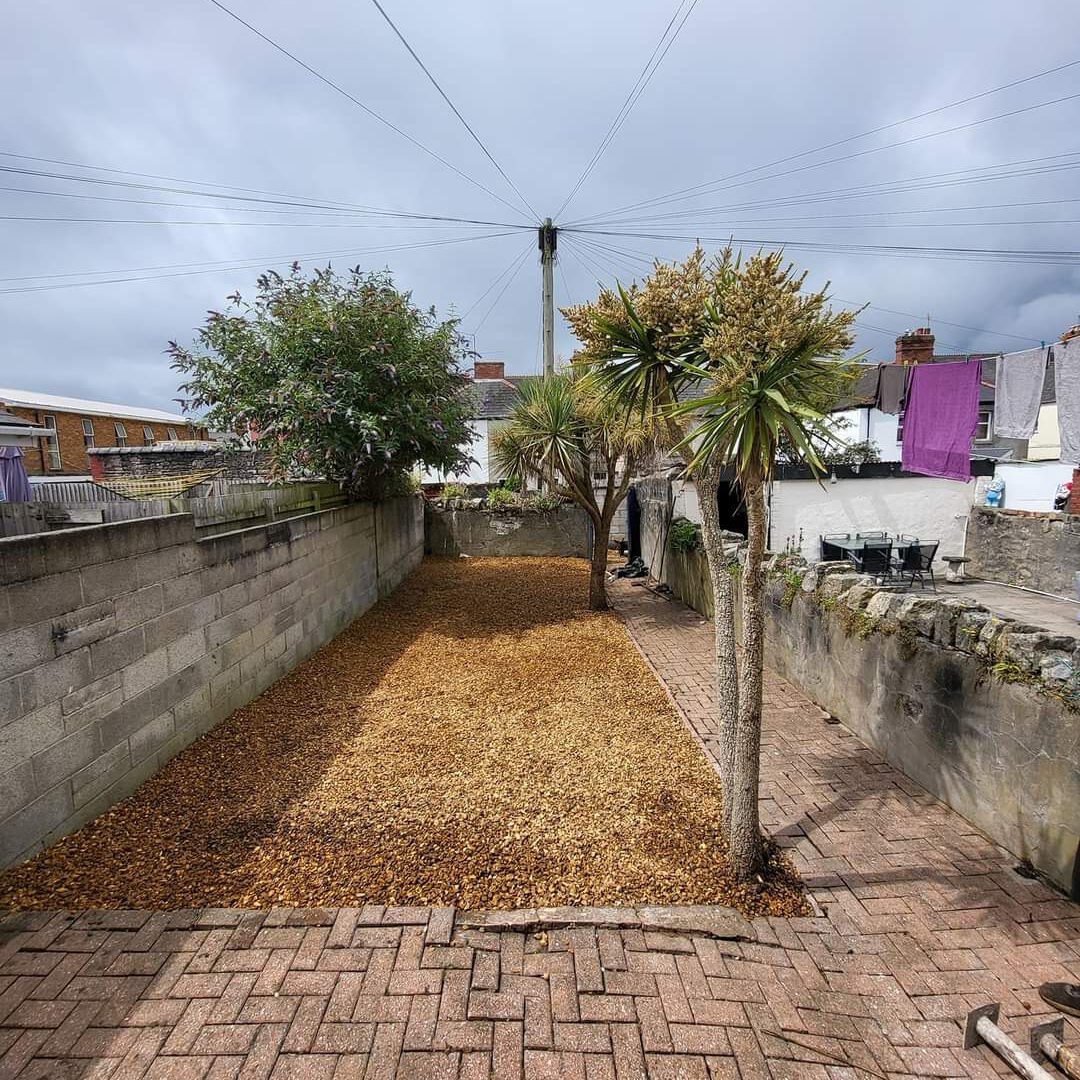 A backyard with a brick-paved area leading to a section covered with wood chips. Two palm trees and a bush are situated along the right side, adjacent to a concrete wall. In the background, there are houses with laundry hanging outside, and the sky is overcast.