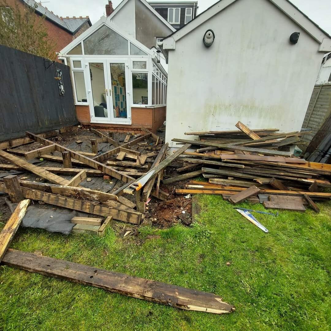 A backyard with dismantled wood decking in disarray. The structure is partially deconstructed, with wooden planks and beams scattered on the ground. A white house with a glass conservatory is in the background. The lawn is green and slightly overgrown.