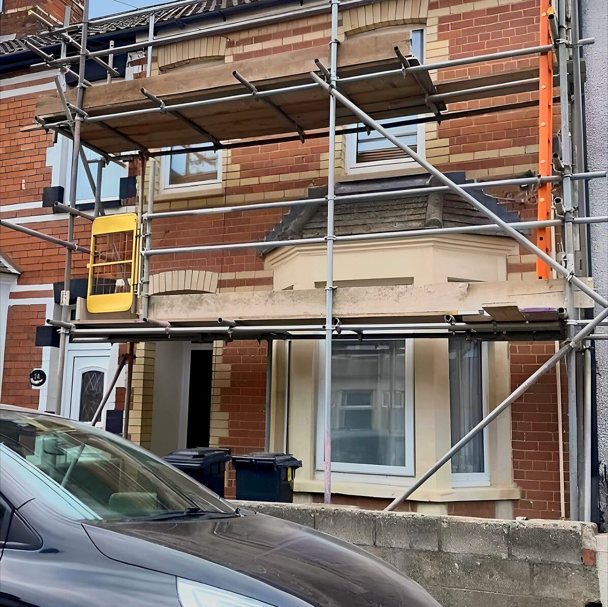 A brick house with white framed windows is under renovation, partially covered with scaffolding. A yellow ladder is situated in the scaffolding, and a black car is parked in front of the house.