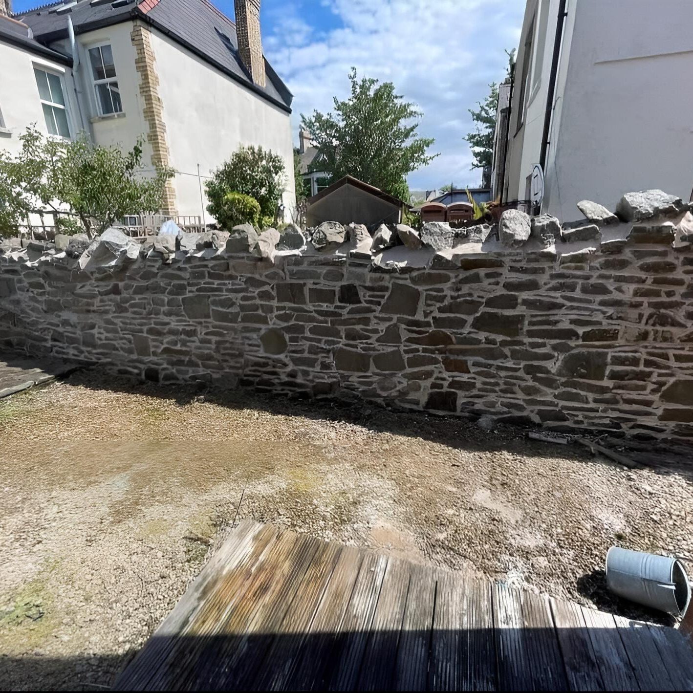 A backyard enclosed by a stone wall, with two adjacent white houses visible in the background. The ground has a mix of gravel and wooden planks, and a small garden shed can be seen beyond the wall. A metal bucket sits in the lower right corner.