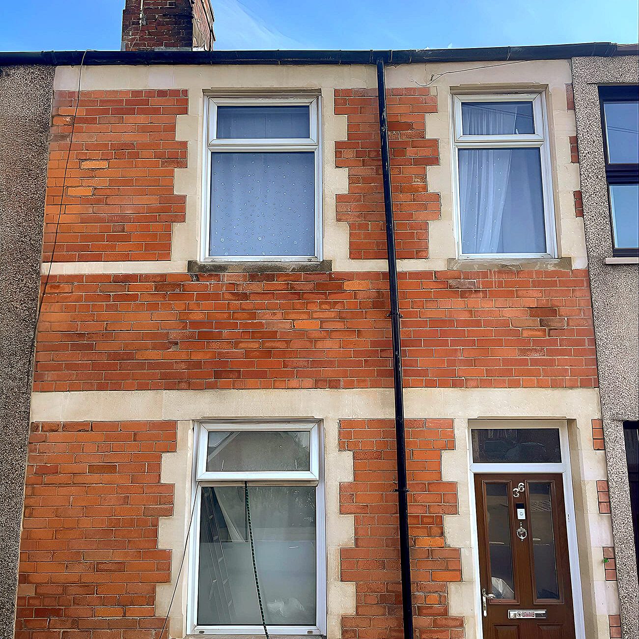 A two-story terraced house with a red brick facade and white trim. It features two upper windows, one lower window, and a dark brown door with a small window at the top. A house number and a doorbell are visible on the door.