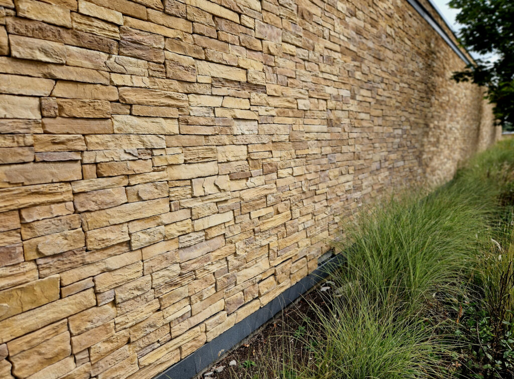 A long stone wall made of rectangular tan bricks stretches into the distance, with tall green grass growing alongside its base. The wall is slightly angled, showing depth and perspective.