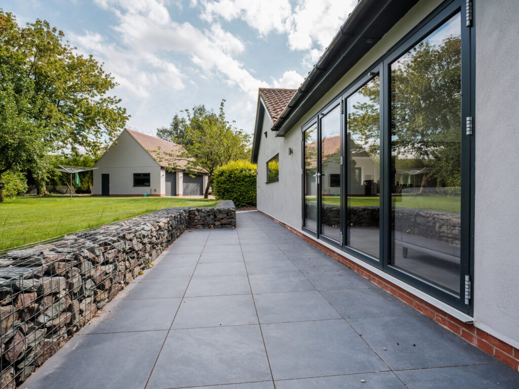 A modern patio with large gray tiles, glass sliding doors, and a stone gabion wall, overlooking a green lawn and another house in the background surrounded by trees.