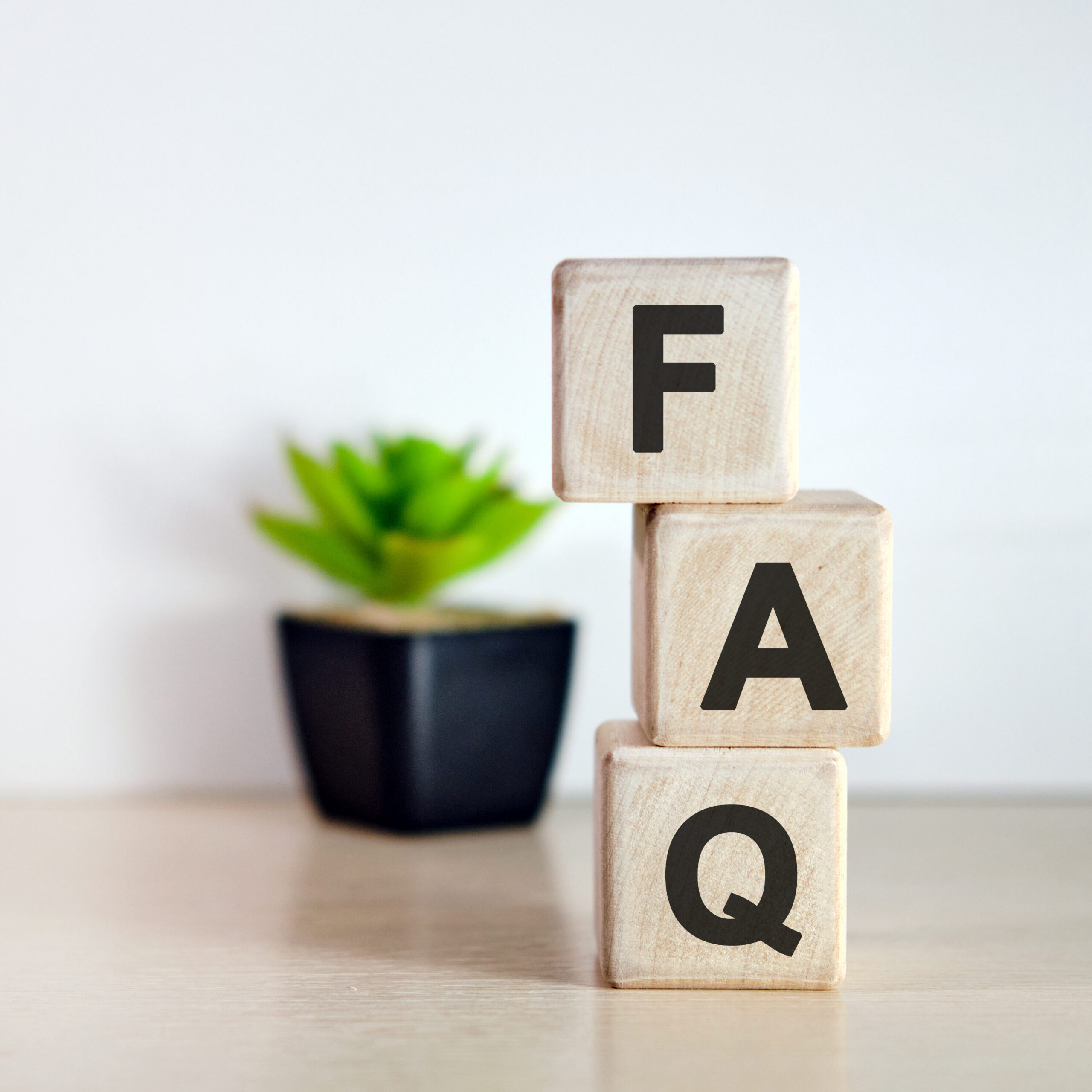 Three wooden blocks stacked vertically with the letters "F", "A", and "Q" in black, spelling "FAQ". A small green potted plant sits in the background on a light-colored surface.