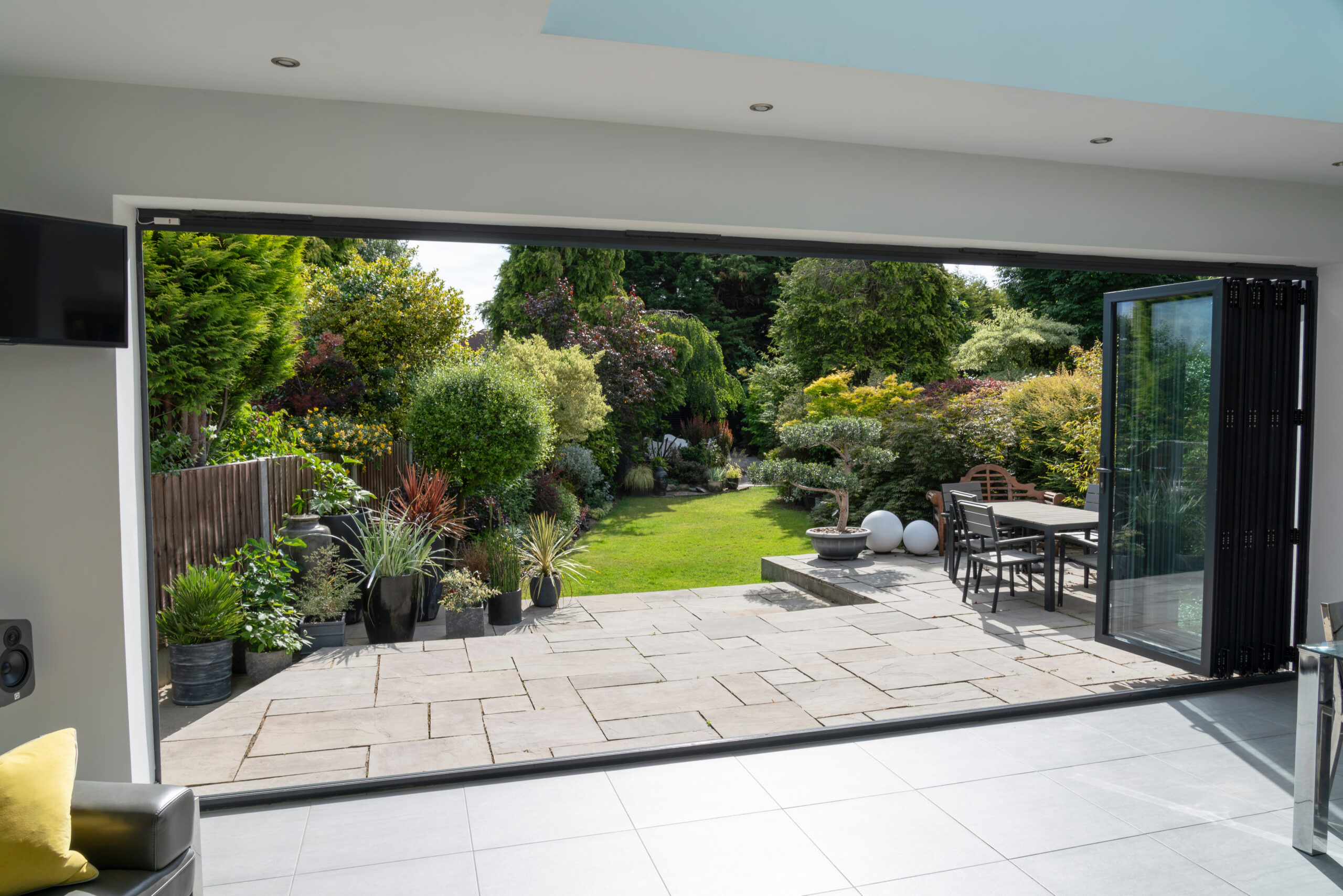 View from inside a modern home looking out through large glass doors onto a stone patio with outdoor dining set, surrounded by lush green plants and garden landscaping.