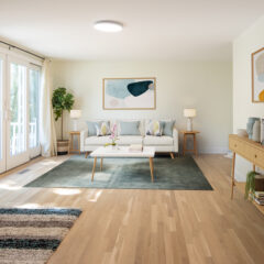 Bright living room with light wood floors, a white sofa, modern art, a green rug, coffee table, sideboard with plants and decor, large windows with curtains, and natural light streaming in.
